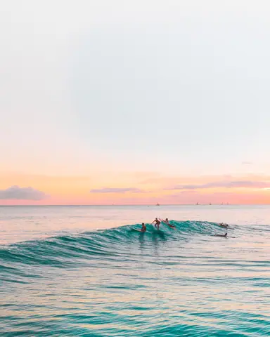 Surfers riding waves at sunset, capturing the serene beauty of the ocean with a pastel sky.