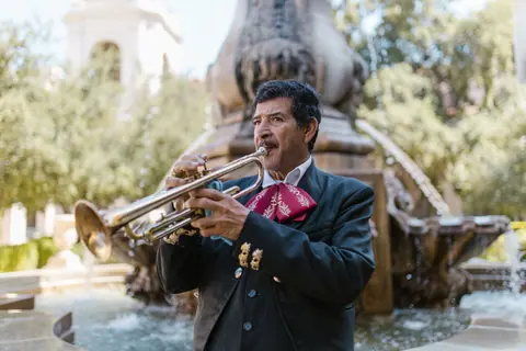 A mariachi musician performs with a trumpet in a lively outdoor setting near a fountain.