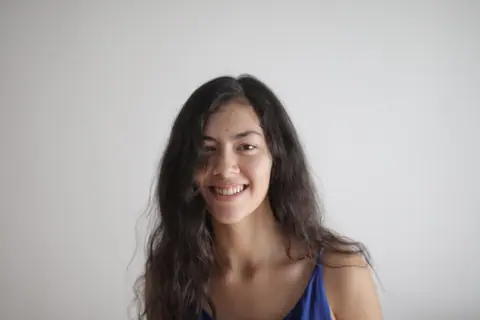 A cheerful woman with long hair smiles warmly in a blue top against a white background.