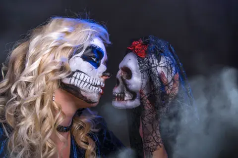 A woman with skull face paint gazes at a veiled skull, creating a spooky Halloween theme.