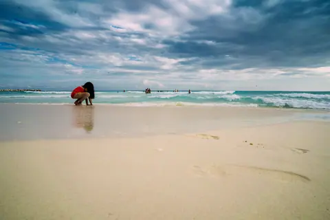 A child in red shorts plays on a sandy tropical beach with a cloudy sky overhead.