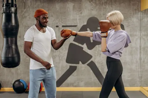 A female athlete spars with her coach in a gym, practicing boxing techniques indoors.