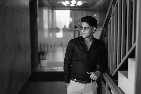 Black and white photo of a stylish adult man leaning against a staircase indoors.