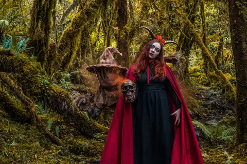 A woman in a Halloween costume holding a skull, standing in a mystical forest.
