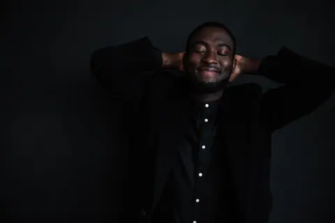 Cheerful young African American male in trendy black shirt with white buttons and black jacket with hands behind head on black background with closed eyes