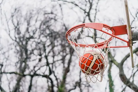 A basketball making a perfect shot through an outdoor hoop against bare winter trees.