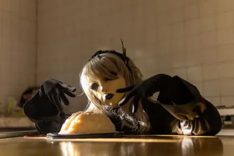 A person in a doll mask and costume poses eerily with a brain-shaped cake, evoking a Halloween theme.