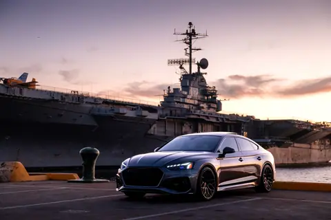 A sleek luxury car parked near a warship at Alameda Harbor with a beautiful sunset backdrop.