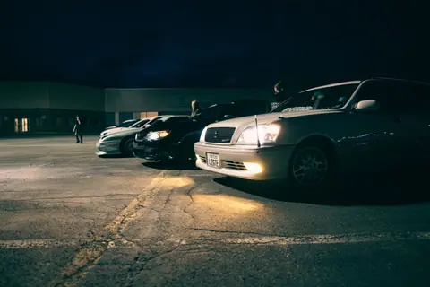 A lineup of cars under parking lot lights during a nighttime car meet.