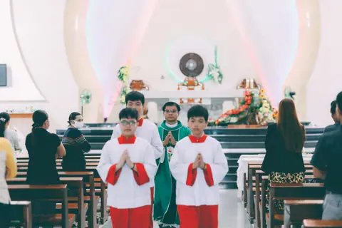 A priest leading altar servers during a church ceremony with congregation present.