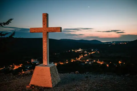 Picturesque sunset with a cross on a hilltop overlooking a peaceful village landscape.