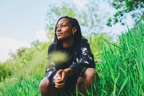 A young woman in stylish streetwear sits crouched in a lush green field, enjoying the outdoors.