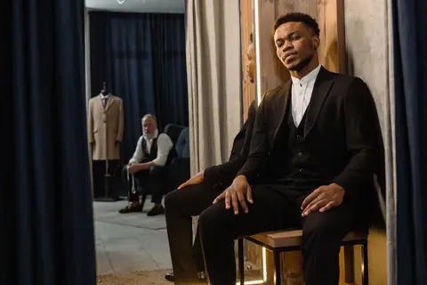African American man in a stylish suit sitting in a modern tailor shop with a mirror reflection.