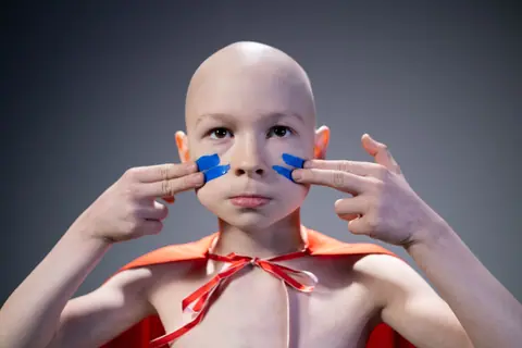 Portrait of a bald child wearing a red cape and blue face paint, embodying strength and courage.
