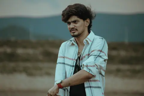 A young man in a plaid shirt poses outdoors with a casual expression.