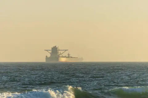 A cargo ship silhouetted against the horizon on a calm ocean during twilight.