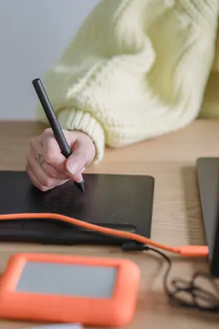 Crop unrecognizable female with stylus drawing on modern black graphic tablet while sitting at wooden table with external hard drive