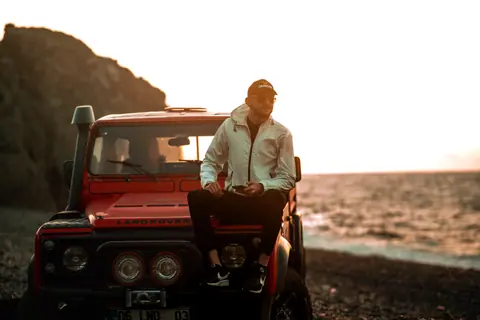 A man sitting on a Land Rover by the beach at sunset, exuding adventure and exploration vibes.