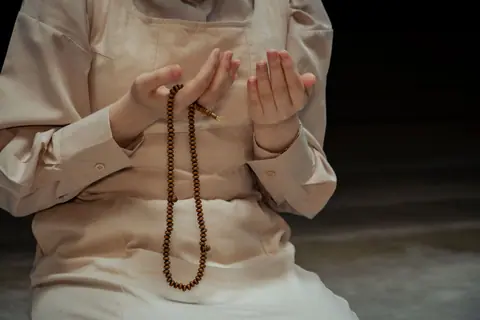 Close-up of an adult praying with a rosary indoors, hands in a prayer position.