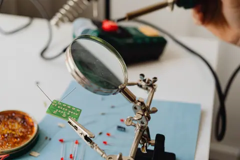 Detailed view of a magnifying glass examining a small circuit board with soldering tools nearby.