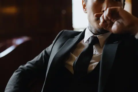 A man in a black suit sitting indoors with a thoughtful expression, hand on face.