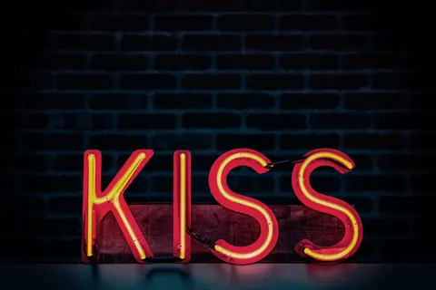 Bright neon lit 'Kiss' sign with a dark brick wall background, ideal for modern decor themes.
