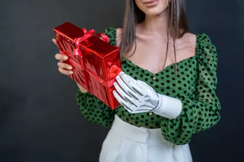 A woman with a prosthetic arm holds a shiny red gift box against a dark background.