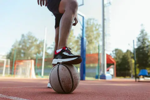 A person standing on an outdoor basketball court with a foot on a basketball, showcasing sports footwear.