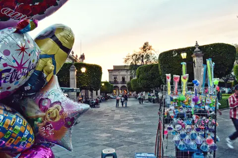 A lively square at sunset featuring colorful balloons, vendors, and a strolling crowd.